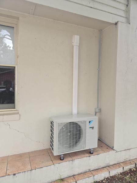 Outdoor condenser beside a rendered wall with a straight, sleeved pipe run to the roof line